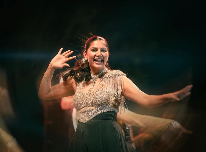 Sapna Choudhary Haryanvi dance performance — TKC Talent booking Sapna Choudhary mid-dance in a silver embellished lehenga on a live stage, smiling with arms open during a Haryanvi dance performance.