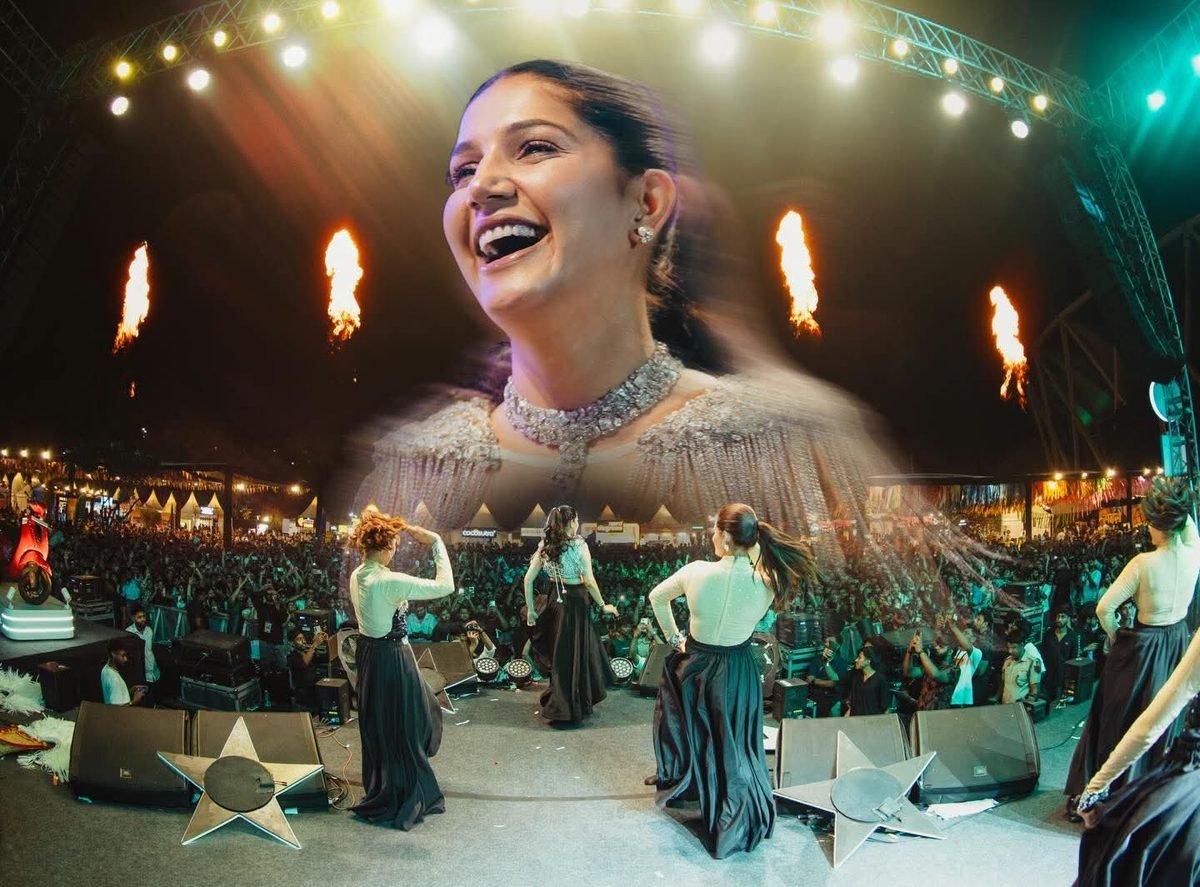 Sapna Choudhary live stage show at night — her face on the giant LED screen with fire jets, dance troupe and a huge crowd at a mela ground.